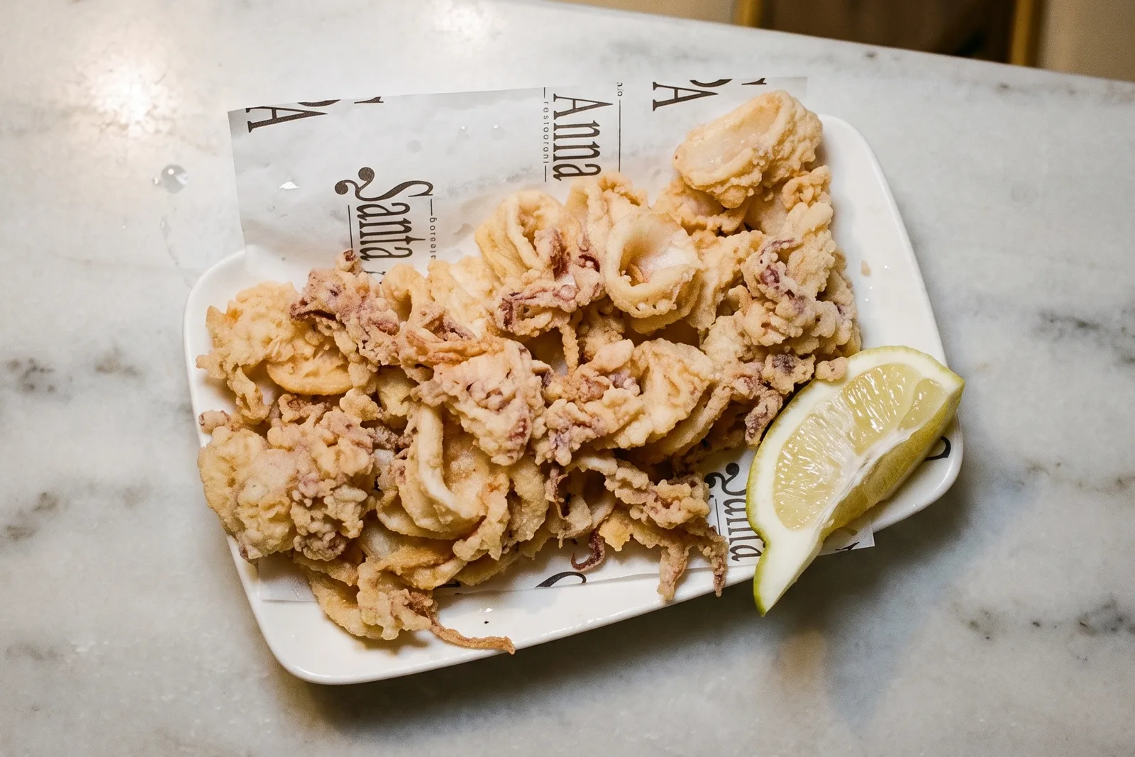Calamares a la andaluza sobre el papel impreso de la casa, con un gajo de limón, en un plato blanco.