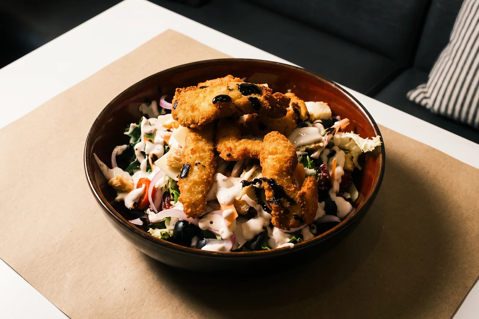 Ensalada César en un bol oscuro, con tiras de pollo rebozado, cebolla morada, aceitunas negras y salsa por encima.