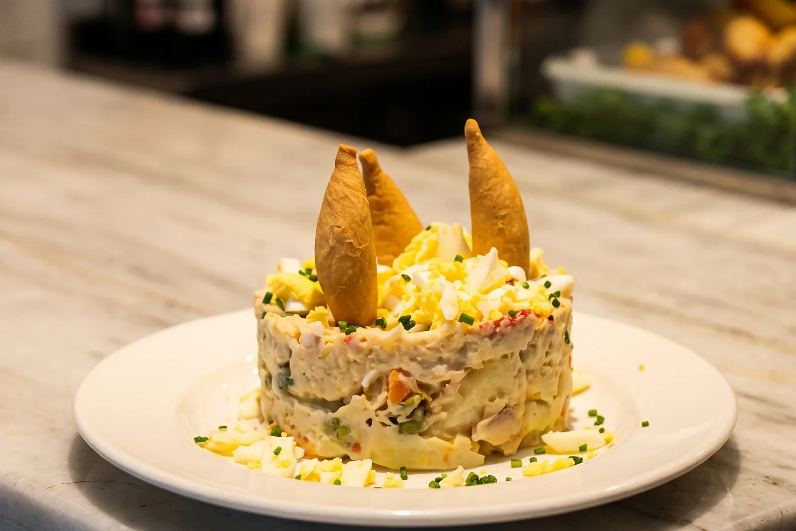 Ensaladilla rusa moldeada en un plato blanco, con huevo duro picado y cebollino alrededor y tres crujientes dorados clavados encima.