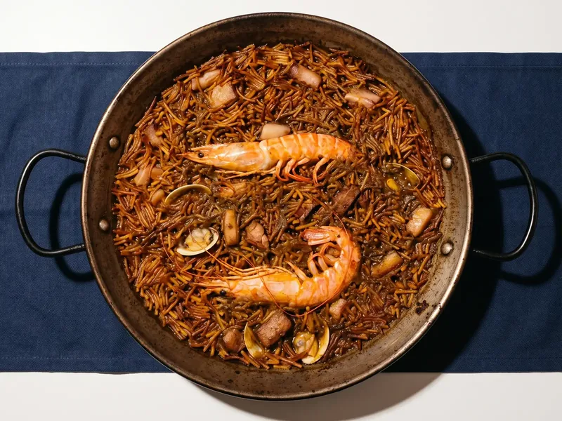 Fideuà en la paellera, con dos gambas enteras, almejas y tacos de carne entre los fideos.