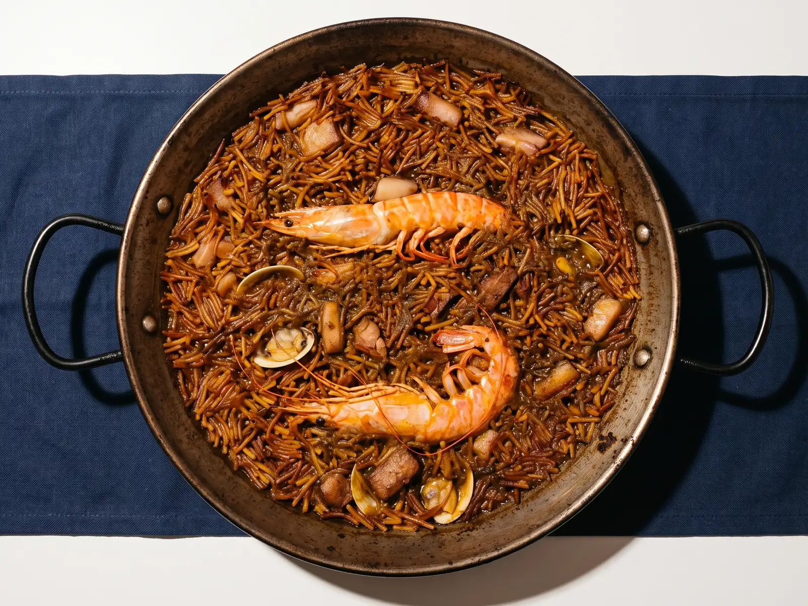 Fideuà en la paellera, con dos gambas enteras, almejas y tacos de carne entre los fideos.