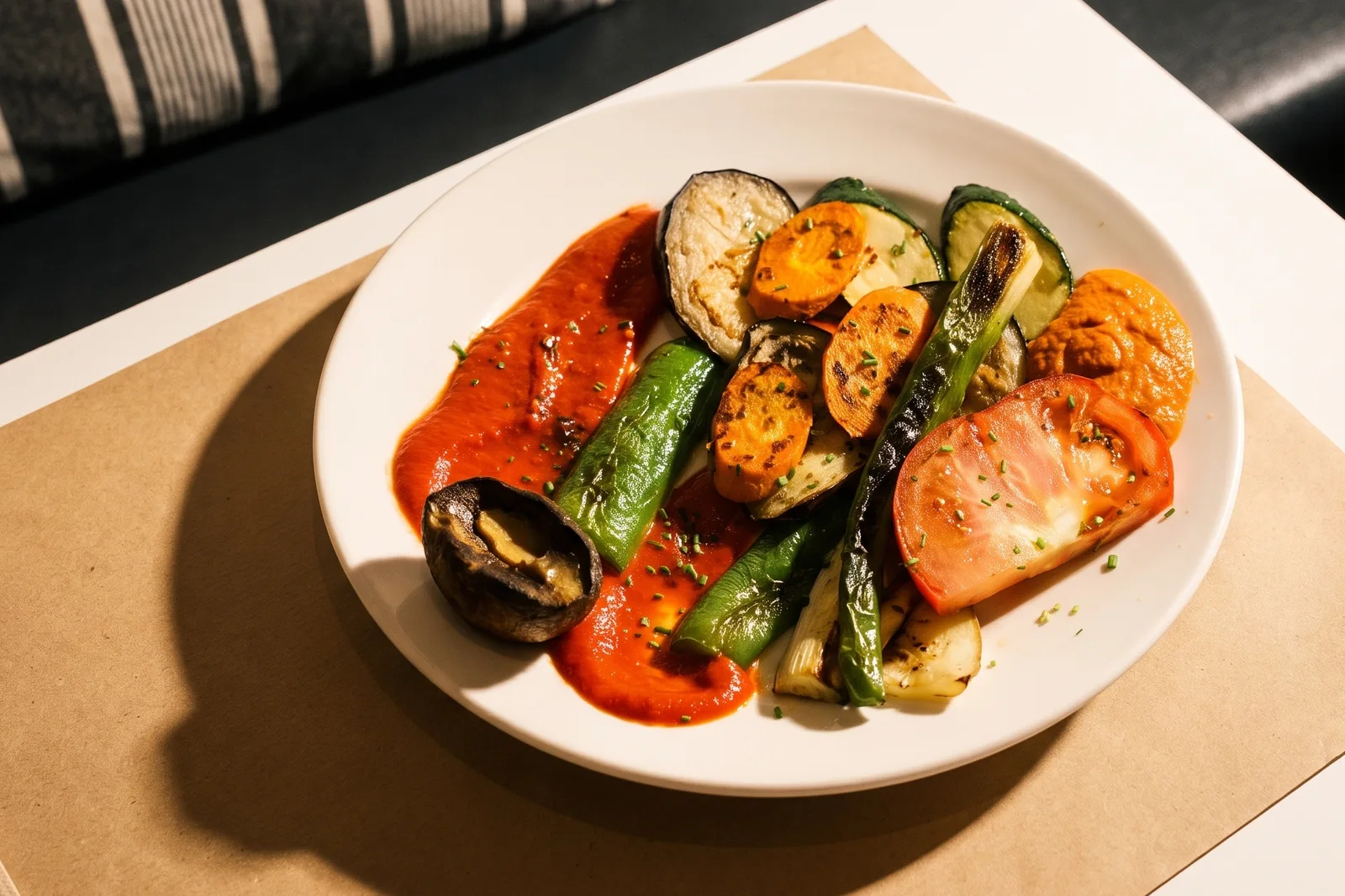 Verduras a la brasa en un plato blanco: berenjena, calabacín, pimiento verde, tomate y champiñón, sobre una salsa roja.
