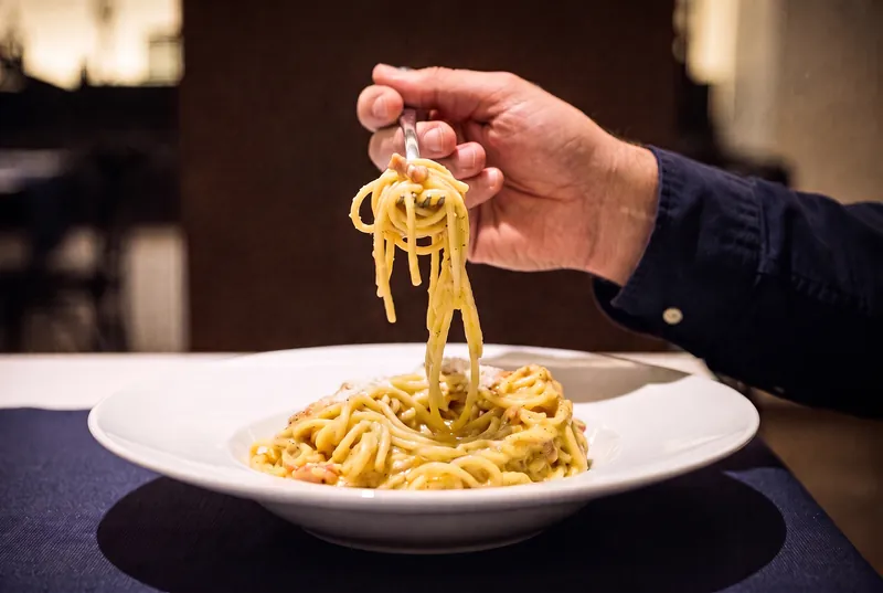 Un tenedor levanta un bocado de espaguetis a la carbonara, enrollados, por encima del plato hondo.