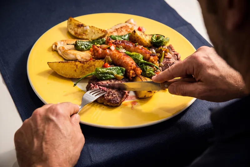 Alguien corta la ternera de una parrillada de carne: butifarra, pechuga de pollo, pimientos verdes y patatas en un plato amarillo.
