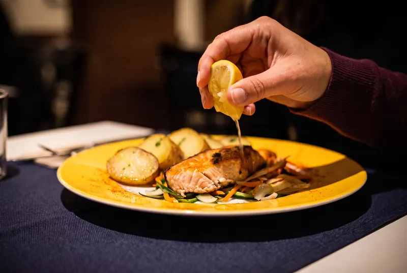 Una mano exprime un gajo de limón sobre un salmón a la plancha, con patatas y verduritas en un plato amarillo.