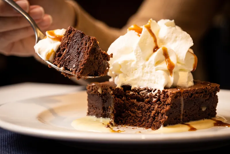 Una cuchara se lleva el primer trozo de una tarta de chocolate con nata montada, caramelo y crema inglesa.