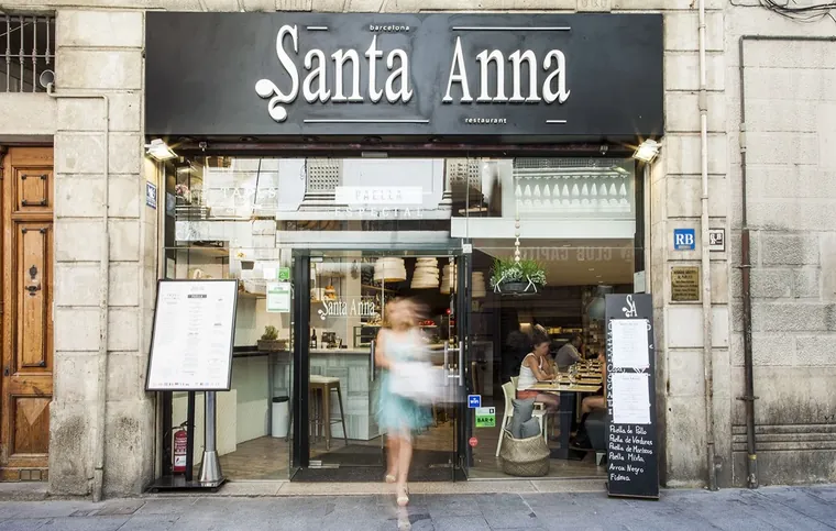 La fachada hoy: el rótulo negro Santa Anna, la sala acristalada con gente comiendo y la pizarra de la acera con las paellas y el arroz negro.