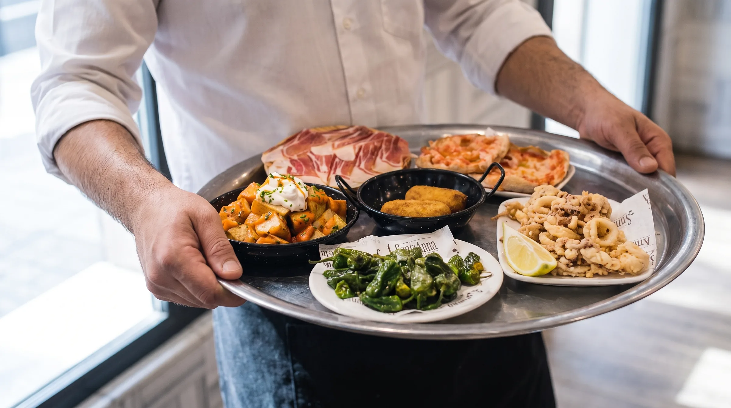 Un camarero lleva una bandeja con seis tapas: patatas bravas, croquetas, pimientos de padrón, pan con tomate, calamares y jamón.