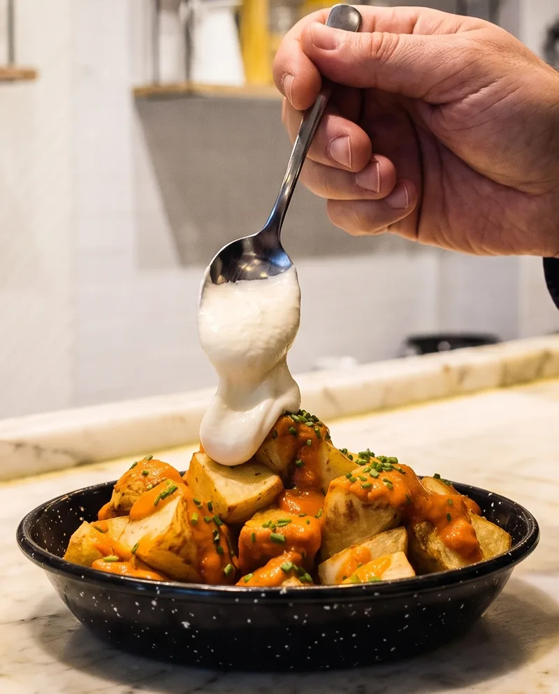 Una cuchara deja caer el alioli sobre una cazuelita de patatas bravas, en la barra de mármol.