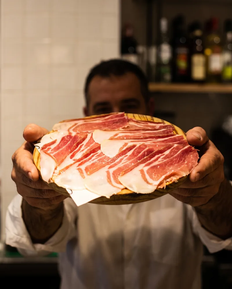 Un camarero acerca la tabla de jamón ibérico, con las botellas de la barra al fondo.