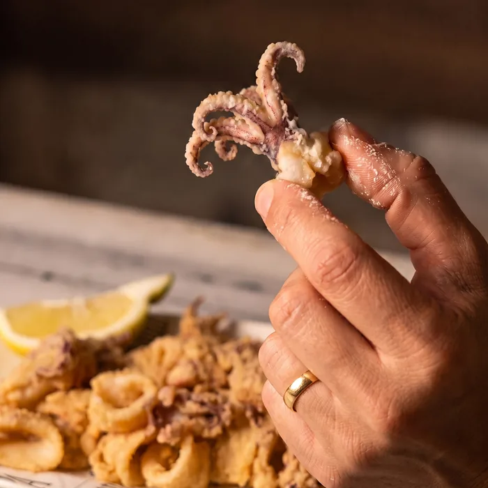 Una mano sostiene un tentáculo de calamar frito, con el plato y el gajo de limón detrás.