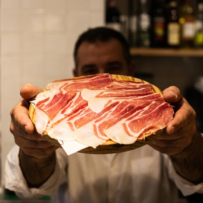 Un camarero acerca la tabla de jamón ibérico, con las botellas de la barra al fondo.