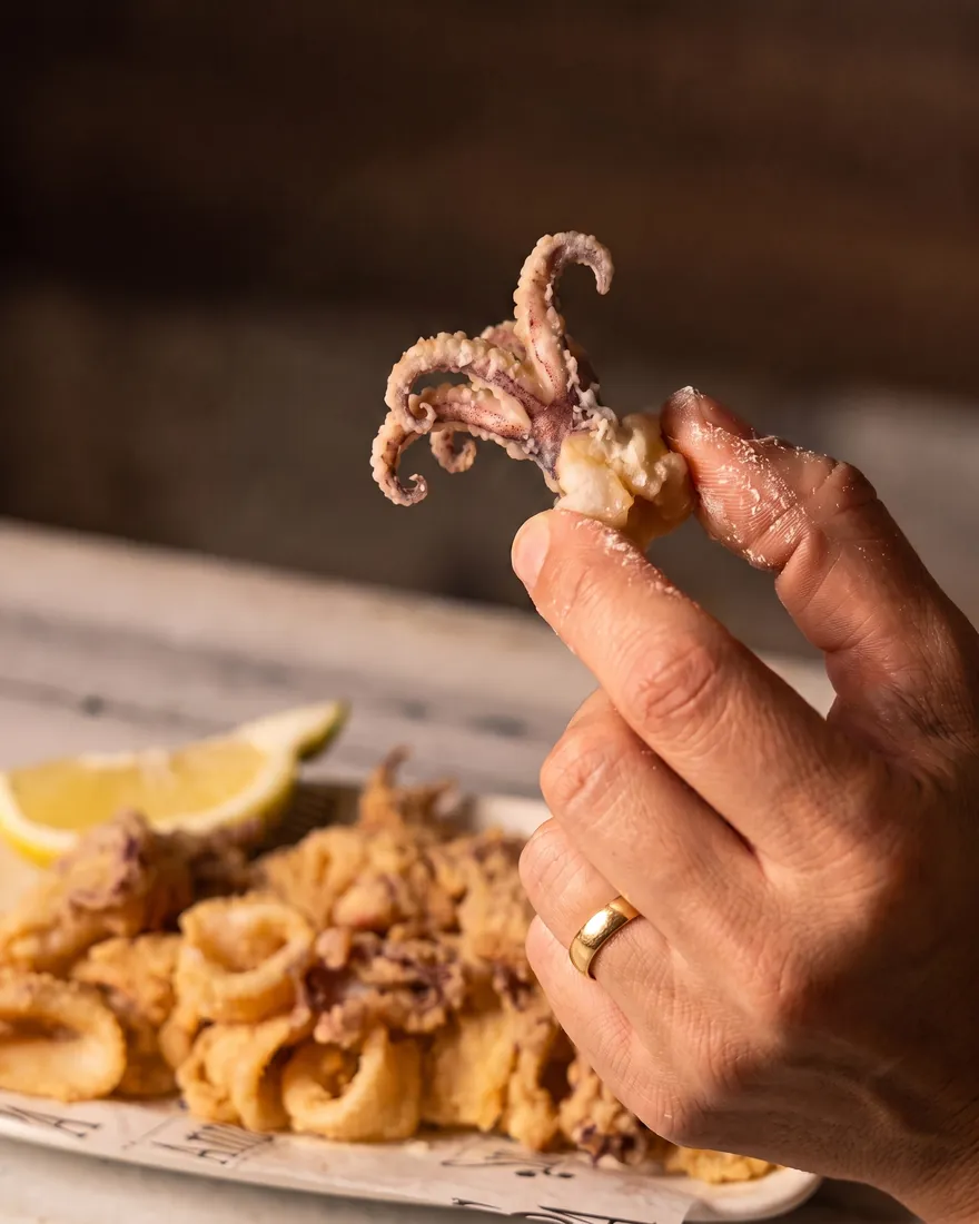 Una mano sostiene un tentáculo de calamar frito, con el plato y el gajo de limón detrás.